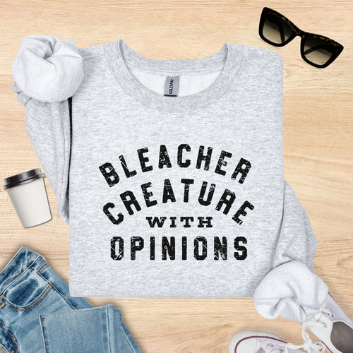 Gray sweatshirt with 'Bleacher Creature with Opinions' text on a wooden surface with accessories.