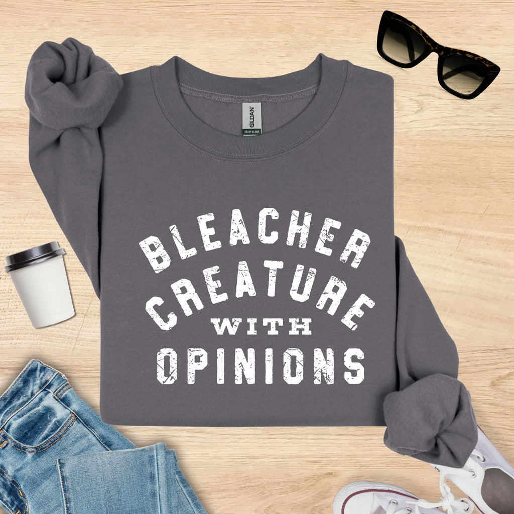 Gray sweatshirt with 'Bleacher Creature with Opinions' text on a wooden surface with sunglasses, jeans, and a cup.