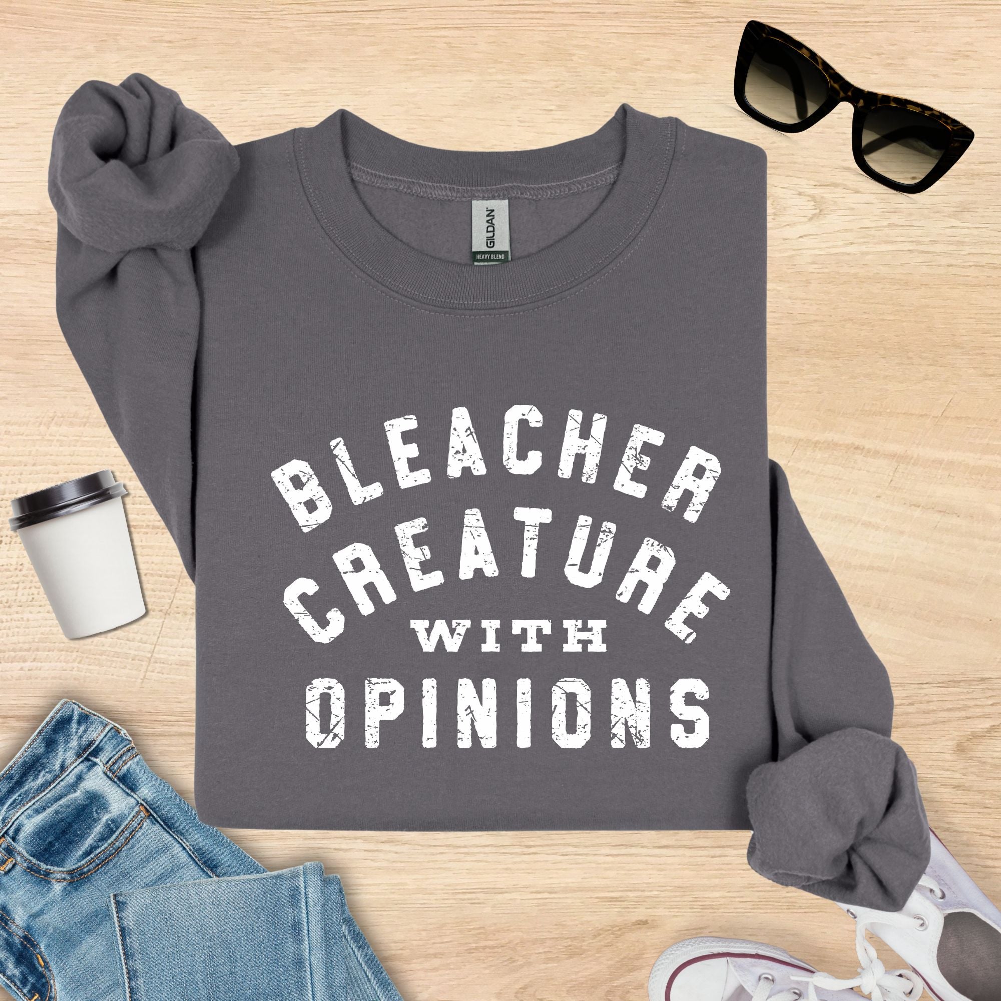 Gray sweatshirt with 'Bleacher Creature with Opinions' text on a wooden surface with sunglasses, jeans, and a cup.