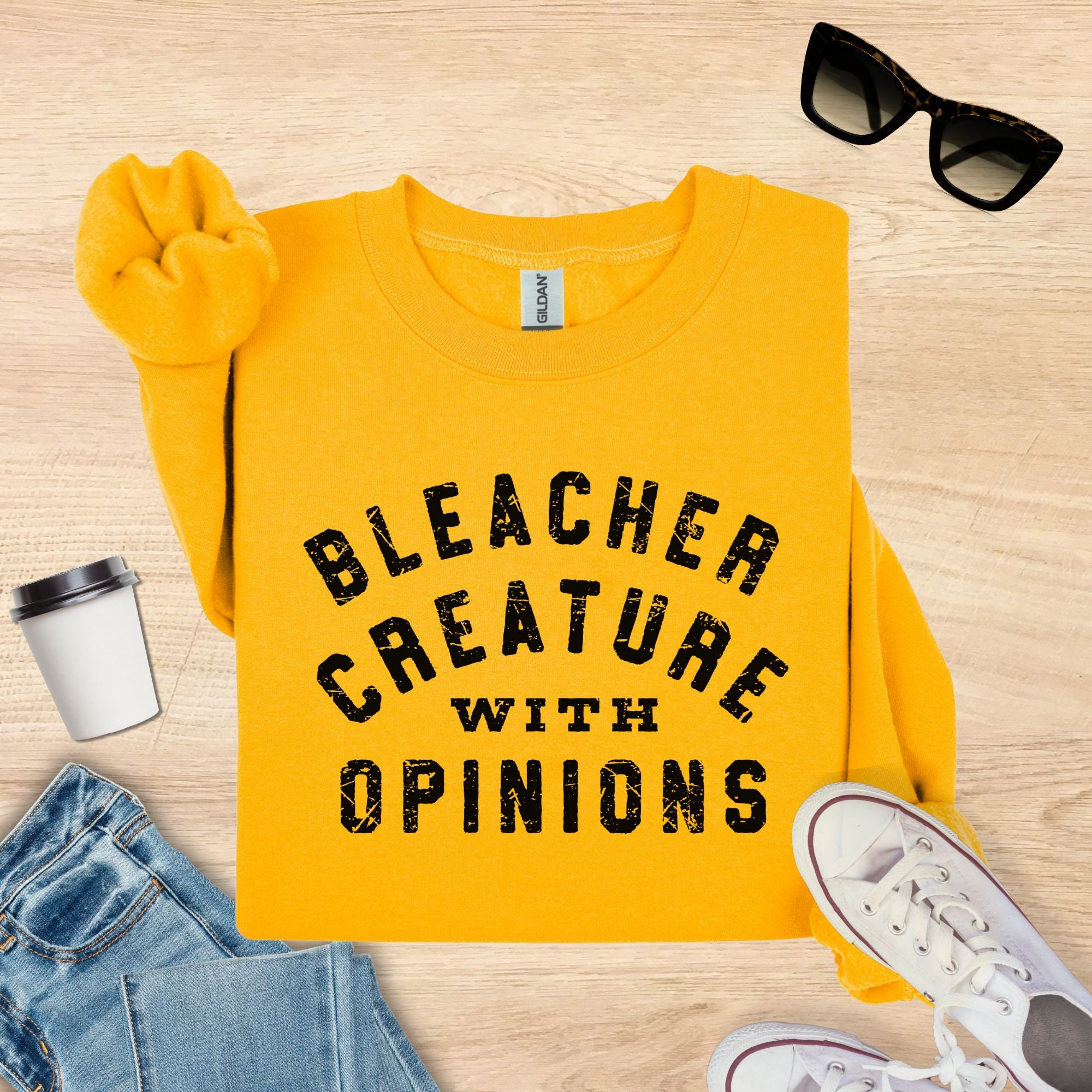 Yellow sweatshirt with 'Bleacher Creature with Opinions' text on a wooden surface with sunglasses, coffee cup, and jeans.