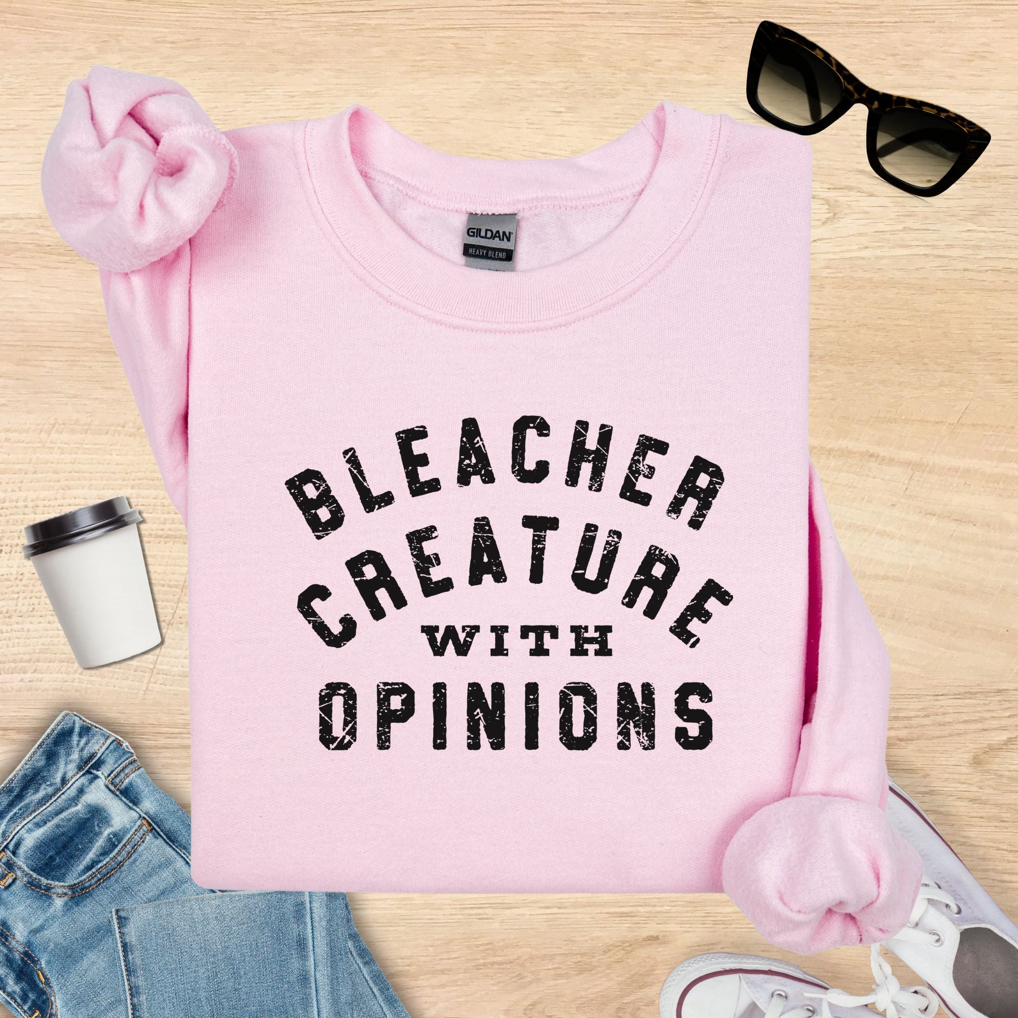 Pink sweatshirt with 'Bleacher Creature with Opinions' text on a wooden surface with accessories.