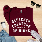 Maroon sweatshirt with 'Bleacher Creature with Opinions' text on a wooden surface with accessories.