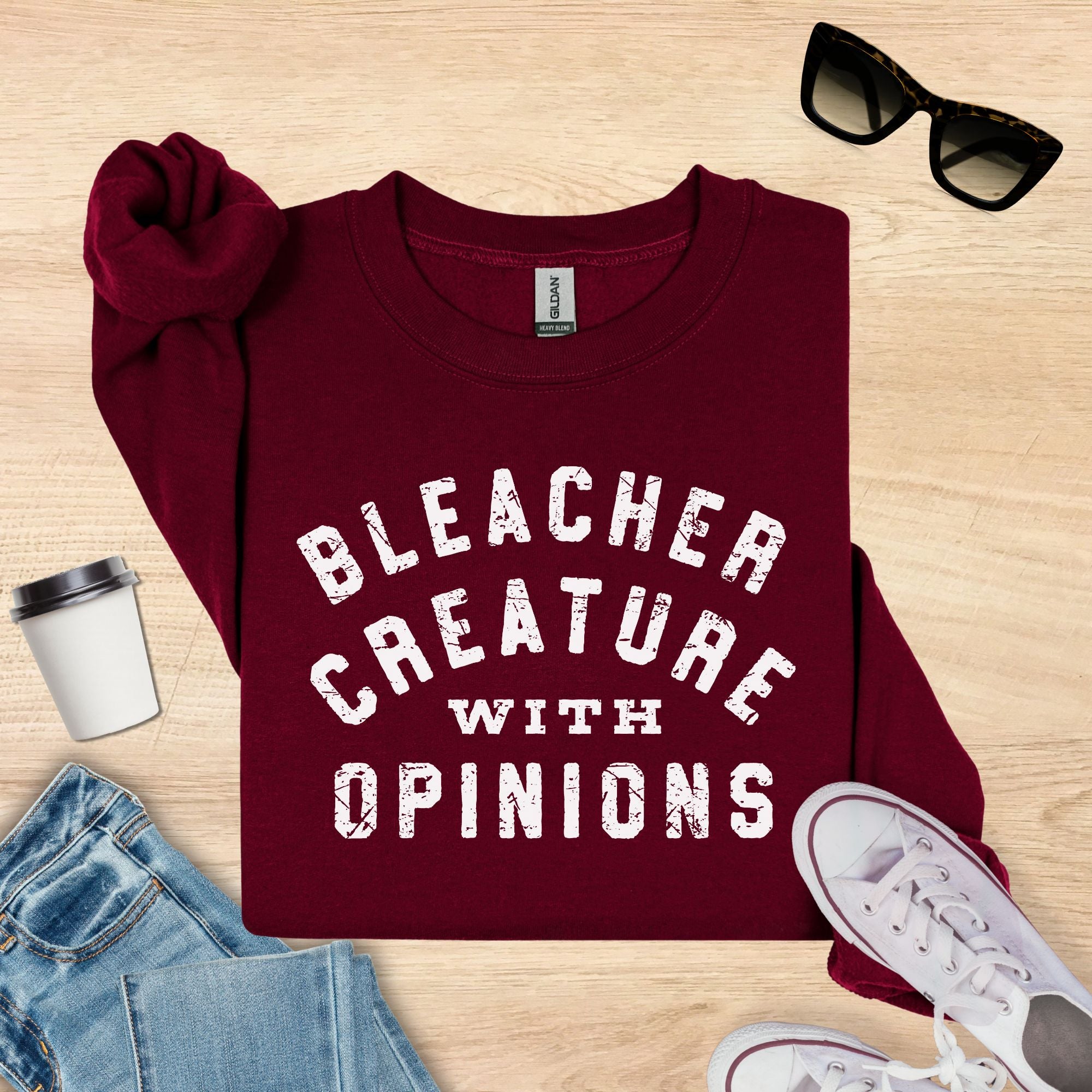Maroon sweatshirt with 'Bleacher Creature with Opinions' text on a wooden surface with accessories.