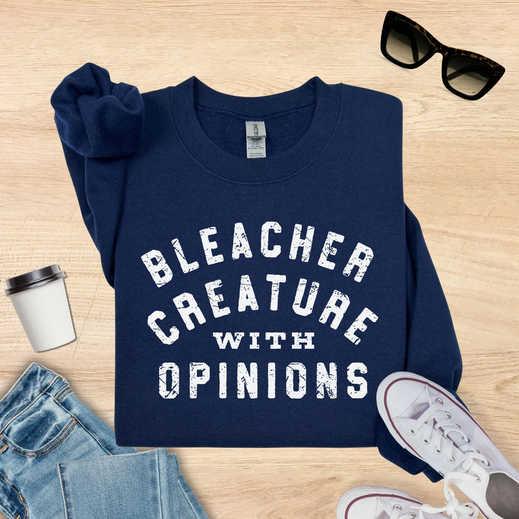 Navy blue sweatshirt with 'Bleacher Creature with Opinions' text, sunglasses, jeans, and sneakers on a wooden surface.