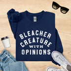Navy blue sweatshirt with 'Bleacher Creature with Opinions' text, sunglasses, jeans, and sneakers on a wooden surface.