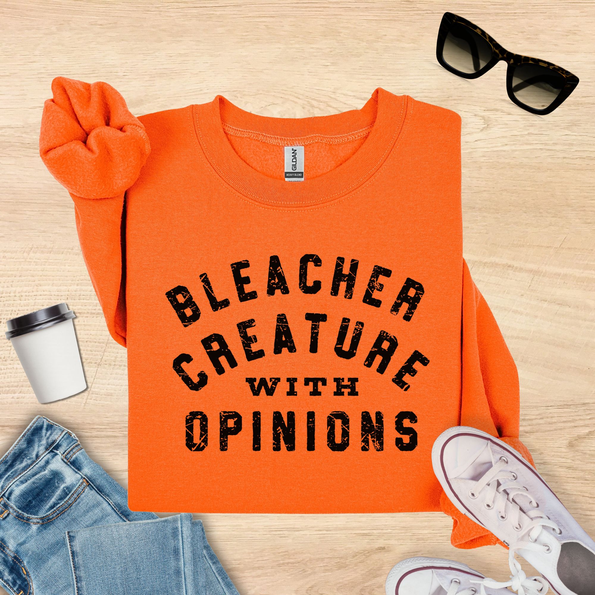 Orange sweatshirt with 'BLEACHER CREATURE WITH OPINIONS' text on a wooden surface with sunglasses, cup, and jeans.