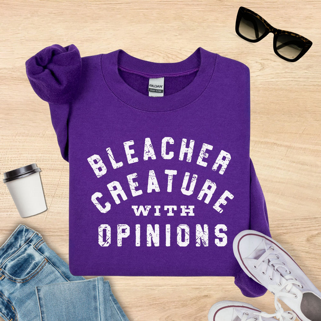 Purple sweatshirt with 'Bleacher Creature with Opinions' text on a wooden surface with sunglasses, coffee cup, and jeans.