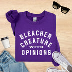 Purple sweatshirt with 'Bleacher Creature with Opinions' text on a wooden surface with sunglasses, coffee cup, and jeans.