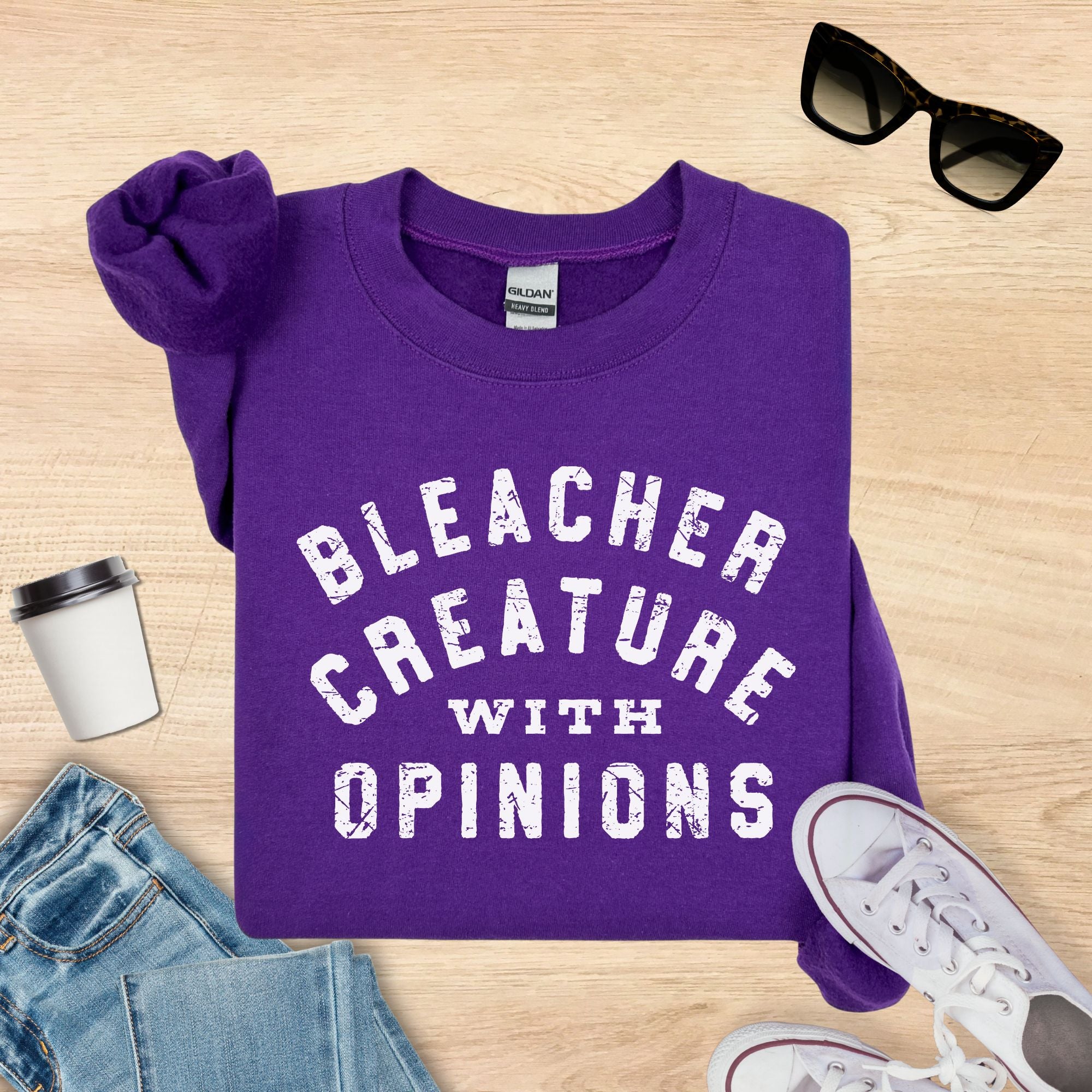 Purple sweatshirt with 'Bleacher Creature with Opinions' text on a wooden surface with sunglasses, coffee cup, and jeans.