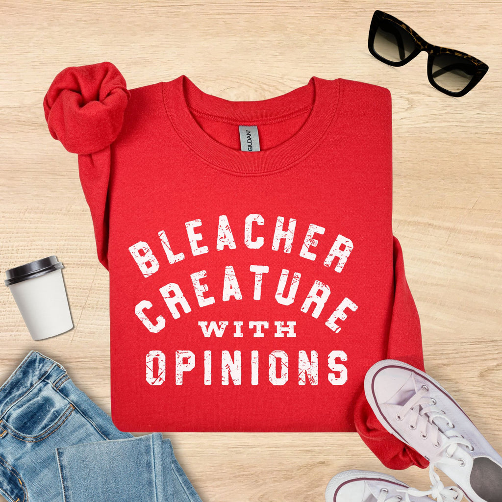 Red t-shirt with 'Bleacher Creature with Opinions' text on a wooden surface with accessories.