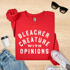 Red t-shirt with 'Bleacher Creature with Opinions' text on a wooden surface with accessories.