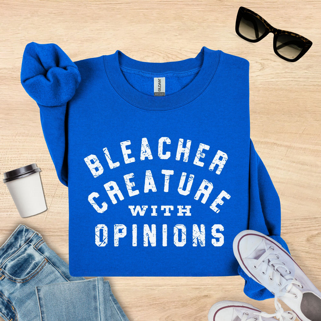 Blue sweatshirt with 'Bleacher Creature with Opinions' text on a wooden surface with sunglasses, cup, and jeans.