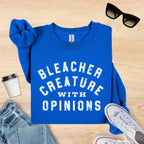 Blue sweatshirt with 'Bleacher Creature with Opinions' text on a wooden surface with sunglasses, cup, and jeans.