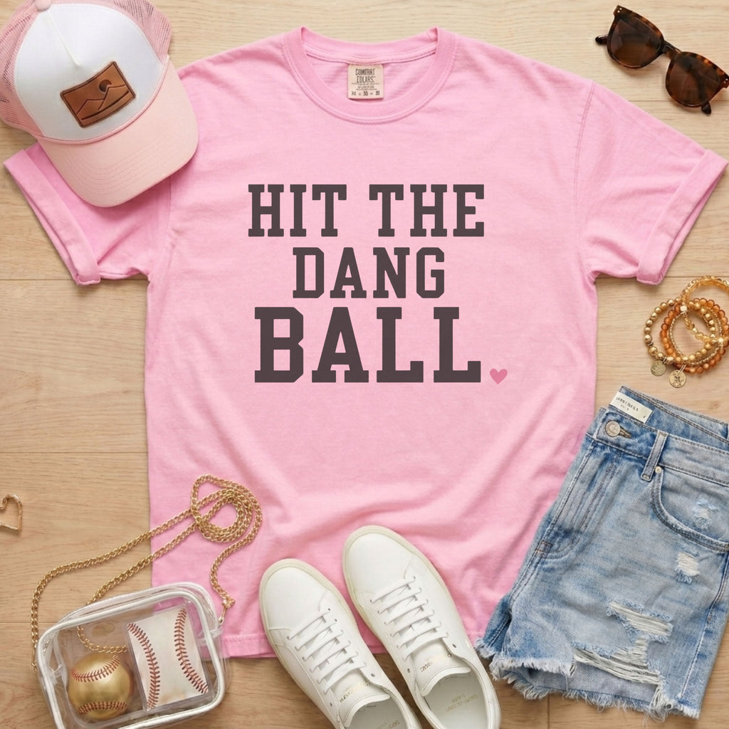 Blossom light pink Comfort Colors Hit The Dang Ball baseball mom shirt styled with denim shorts, sneakers, pink hat, sunglasses, and clear baseball bag.