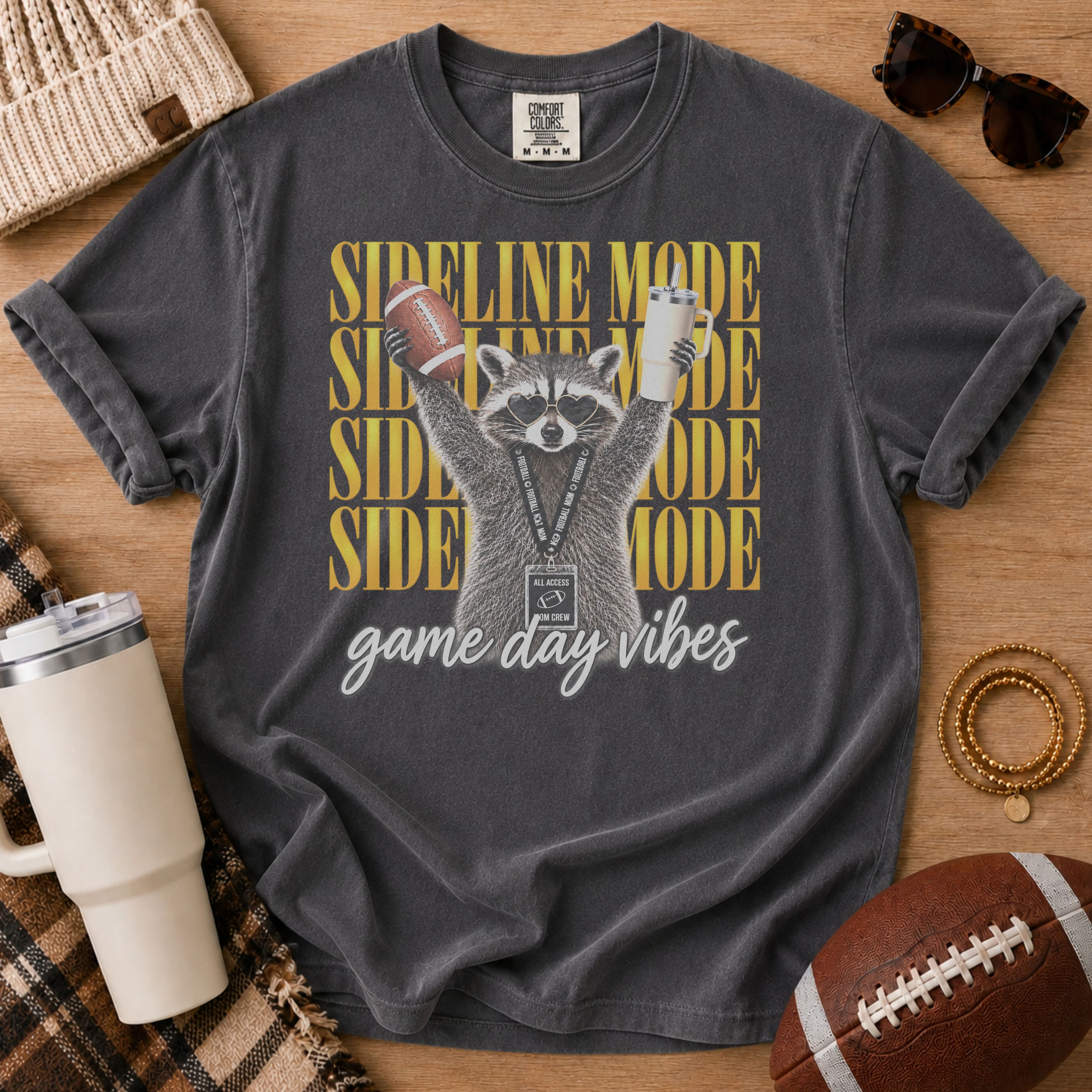 Charcoal gray football mom shirt, Comfort Colors Pepper, styled in a game day flat lay with a football and a raccoon holding a football and tumbler beneath “Sideline Mode” lettering.