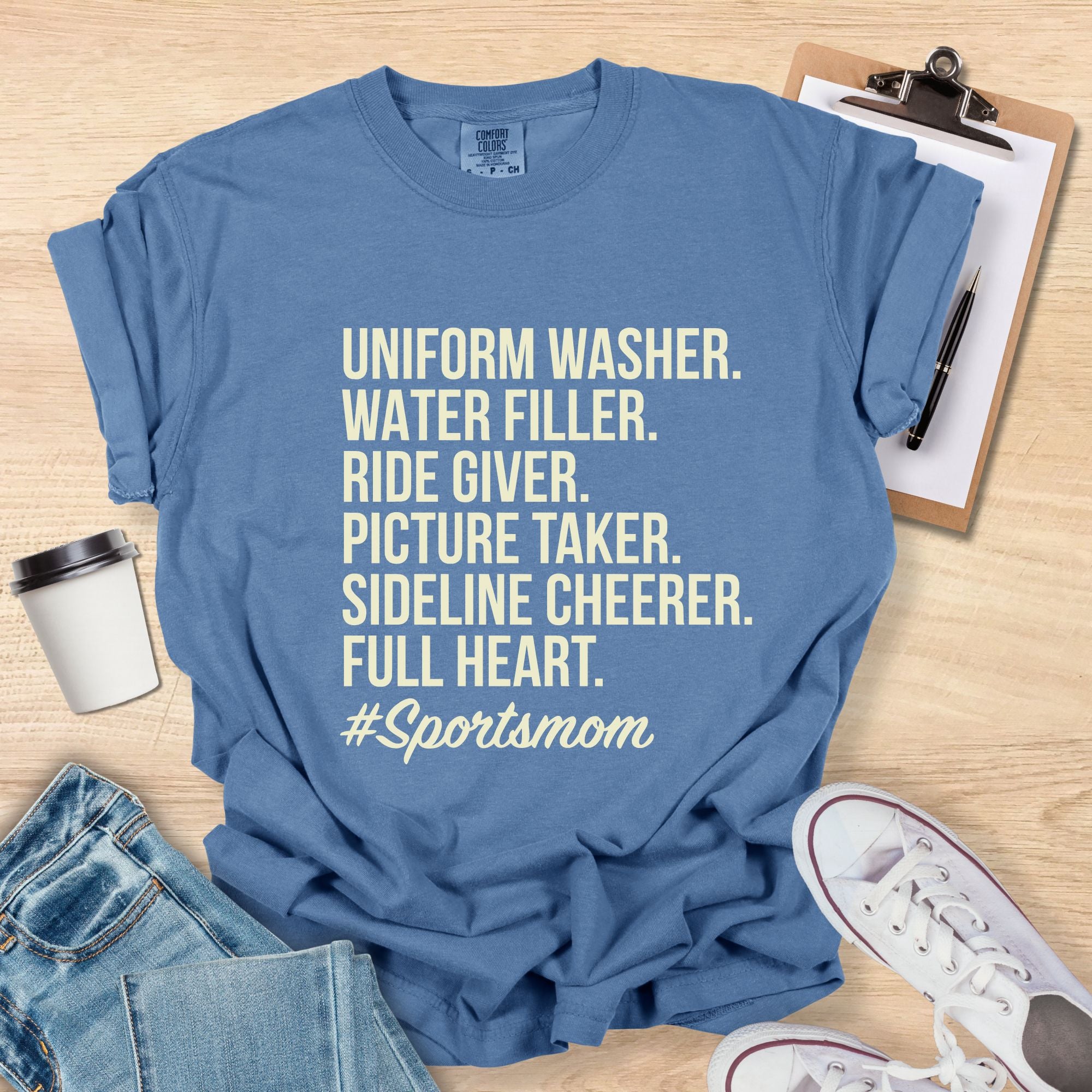 Blue t-shirt with '#Sportsmom' text on a wooden surface with accessories.