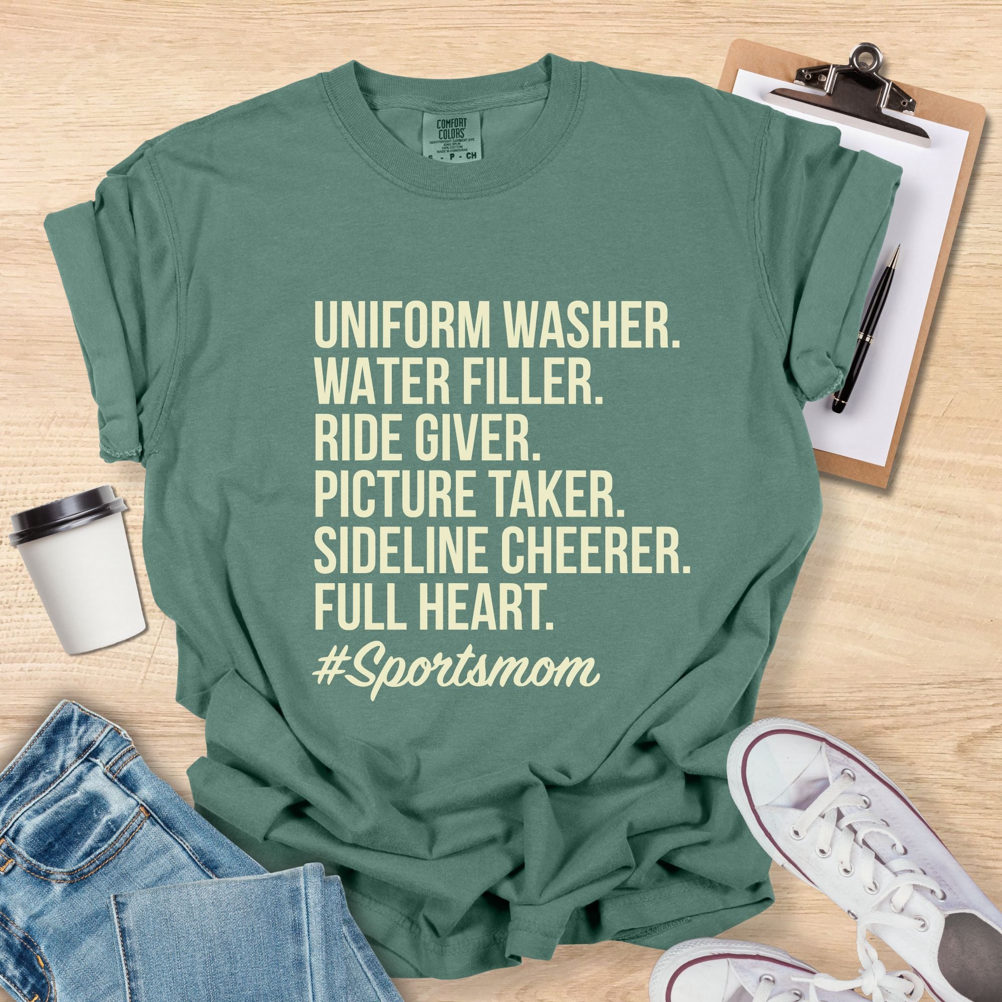 Green t-shirt with '#Sportsmom' text on a wooden surface with accessories.