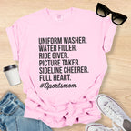 Pink t-shirt with '#Sportsmom' text on a wooden surface with sunglasses, jeans, and sneakers.