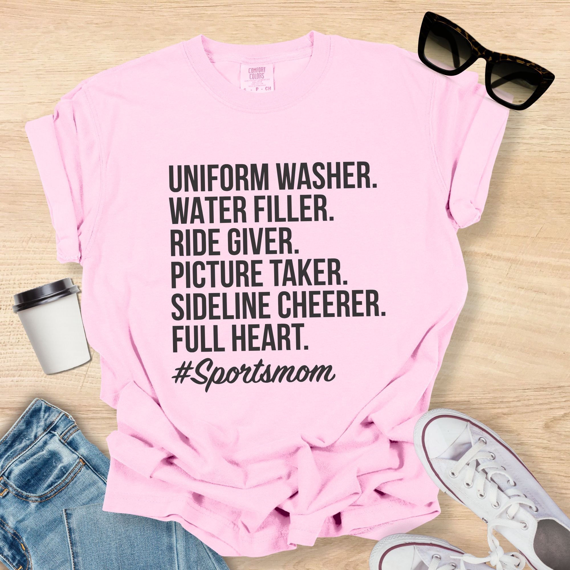 Pink t-shirt with '#Sportsmom' text on a wooden surface with sunglasses, jeans, and sneakers.