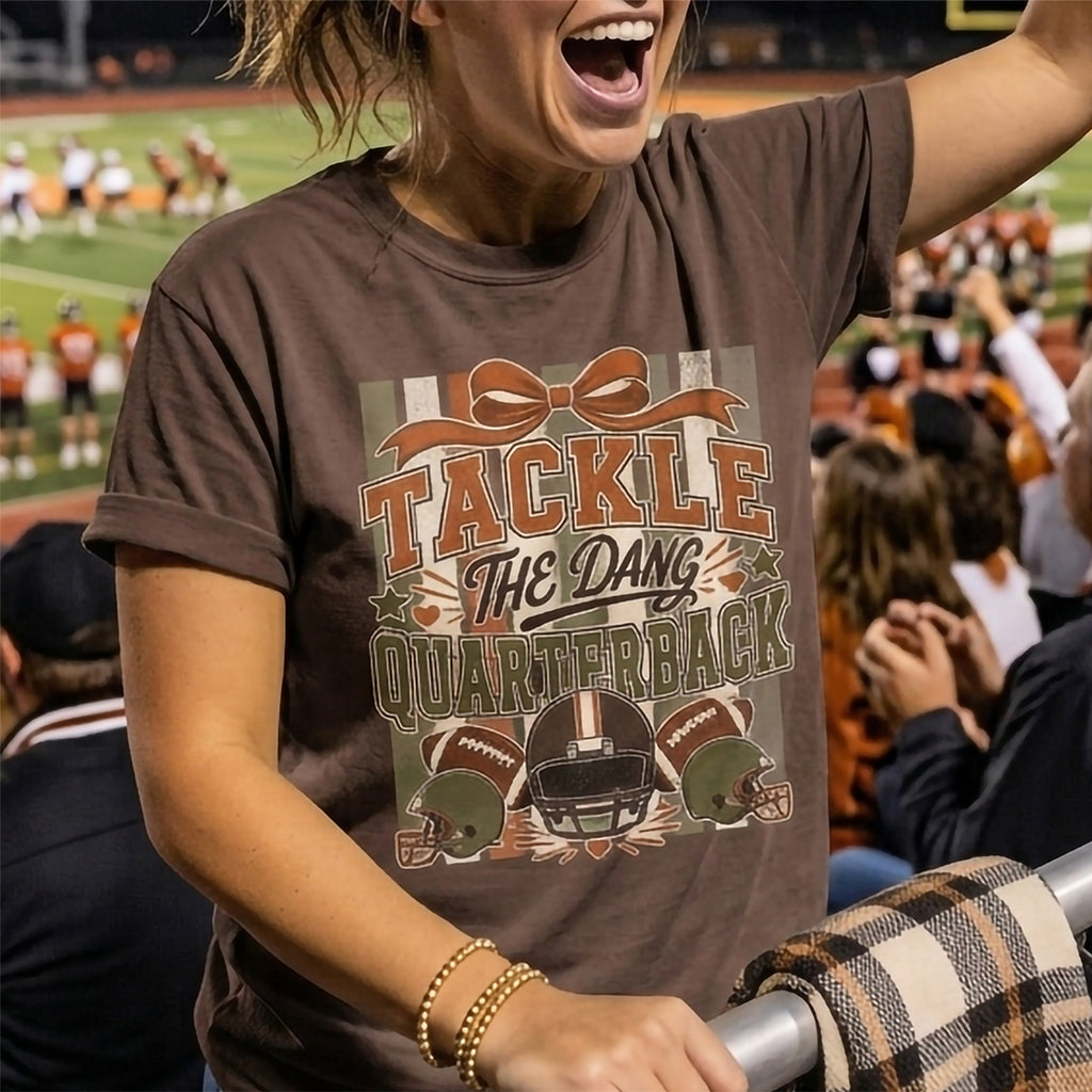 A woman at a football game wearing a brown graphic t-shirt that reads 'TACKLE THE DANG QUARTERBACK' with football helmets and balls, with a blurred stadium crowd in the background.