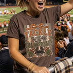 A woman at a football game wearing a brown graphic t-shirt that reads 'TACKLE THE DANG QUARTERBACK' with football helmets and balls, with a blurred stadium crowd in the background.