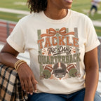 A woman sitting on a track field bleachers wearing a cream-colored graphic t-shirt with a football-themed design that reads 'TACKLE THE DANG QUARTERBACK'. The background shows a blurred football field at sunset.