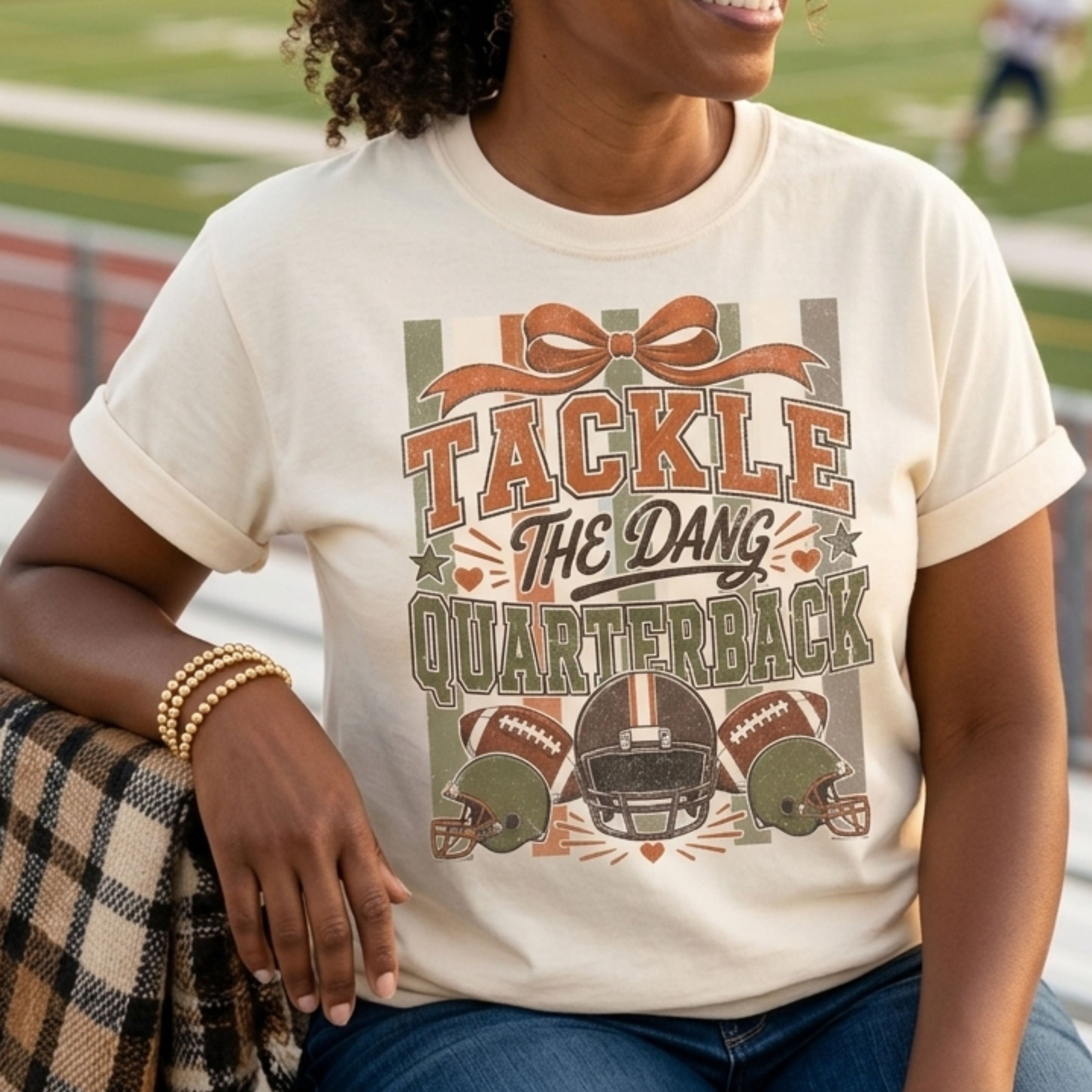 A woman sitting on a track field bleachers wearing a cream-colored graphic t-shirt with a football-themed design that reads 'TACKLE THE DANG QUARTERBACK'. The background shows a blurred football field at sunset.