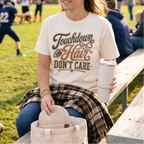 A woman sits on a wooden bench at a football field, wearing a cream graphic T-shirt that reads 'Touchdown Hair DON'T CARE' with a football and heart design. She holds a white insulated tumbler, with a beige knit hat in a tote bag beside her and a plaid flannel shirt draped over her lap.