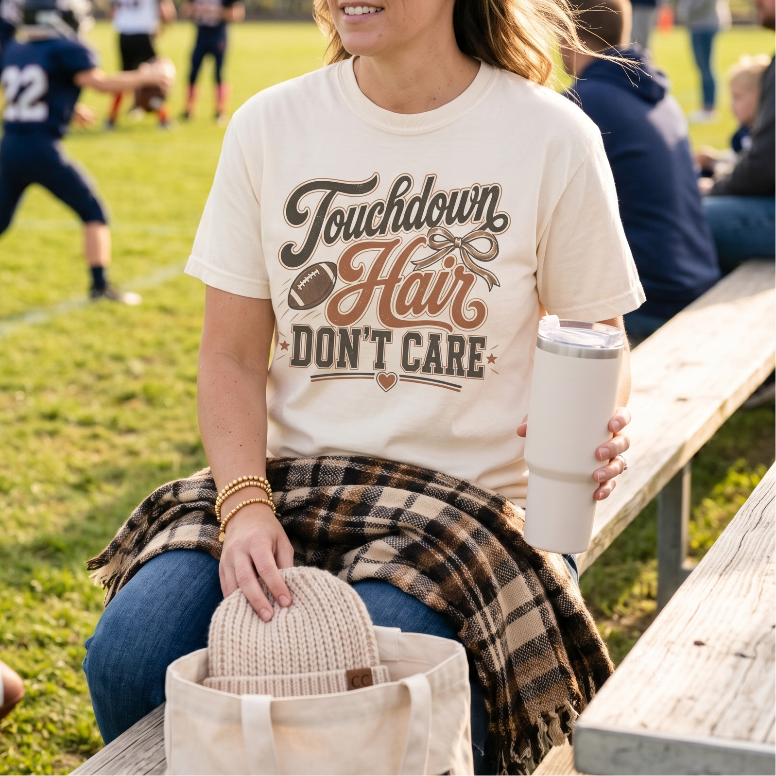 A woman sits on a wooden bench at a football field, wearing a cream graphic T-shirt that reads 'Touchdown Hair DON'T CARE' with a football and heart design. She holds a white insulated tumbler, with a beige knit hat in a tote bag beside her and a plaid flannel shirt draped over her lap.