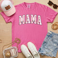 Pink t-shirt with 'MAMA' print, white sneakers, denim shorts, baseball-themed accessories on a wooden surface.