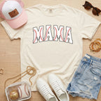 White t-shirt with 'MAMA' print, white sneakers, denim shorts, pink cap, and baseball-themed accessories on a wooden surface.
