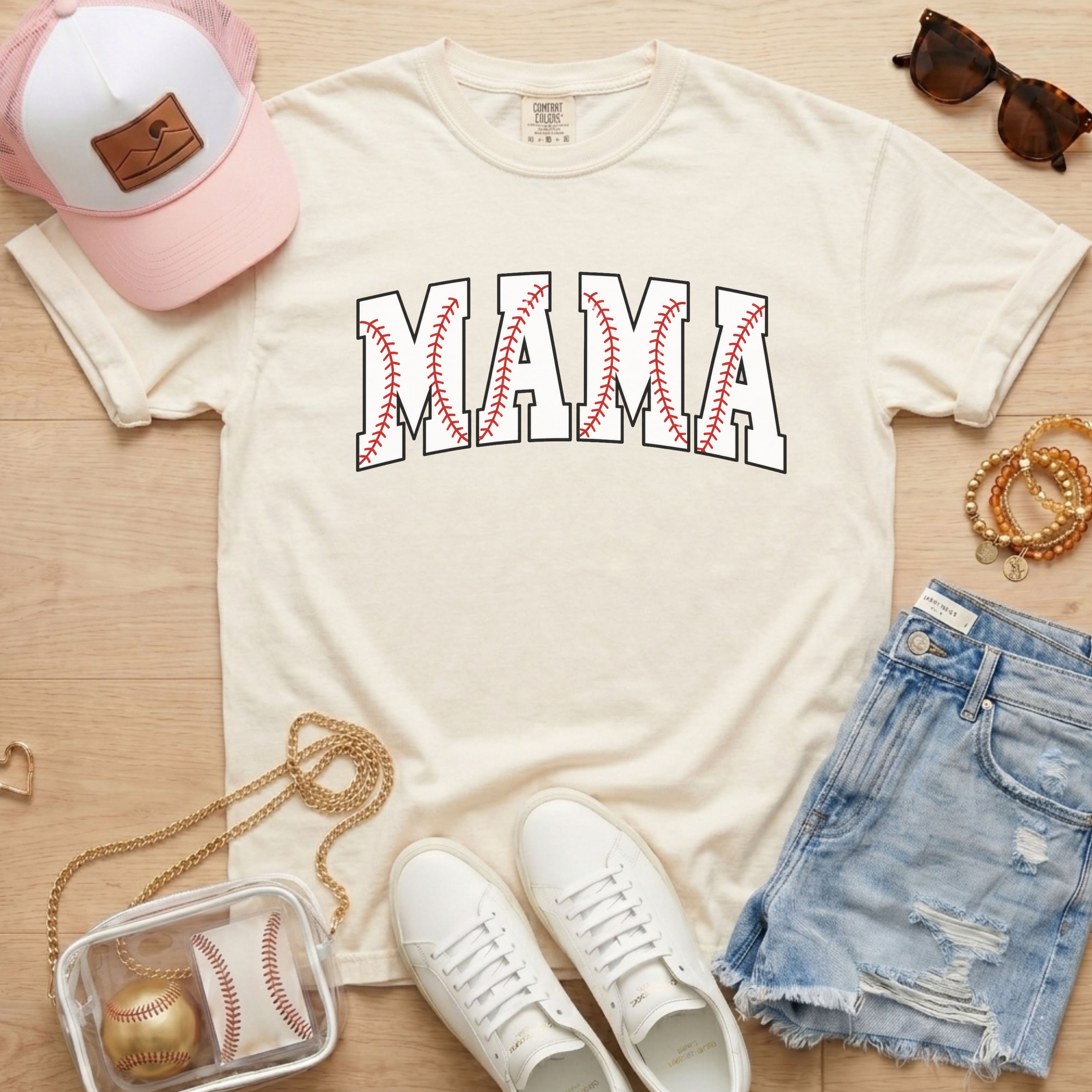 White t-shirt with 'MAMA' print, white sneakers, denim shorts, pink cap, and baseball-themed accessories on a wooden surface.