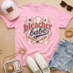 Bleacher Babe baseball mom Comfort Colors tee in blossom light pink flat lay with trucker hat, denim shorts, white sneakers, and baseball.