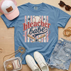 Bleacher Babe baseball mom Comfort Colors tee in blue jean flat lay with trucker hat, denim shorts, white sneakers, and baseball.