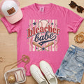 Bleacher Babe baseball mom Comfort Colors tee in crunchberry flat lay with trucker hat, denim shorts, white sneakers, and baseball.
