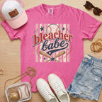 Bleacher Babe baseball mom Comfort Colors tee in crunchberry flat lay with trucker hat, denim shorts, white sneakers, and baseball.