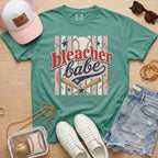 Bleacher Babe baseball mom Comfort Colors tee in light green flat lay with trucker hat, denim shorts, white sneakers, and baseball.