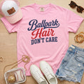 Blossom light pink Comfort Colors Ballpark Hair Don’t Care funny baseball mom shirt styled with denim shorts, sneakers, pink hat, sunglasses, and clear baseball bag.