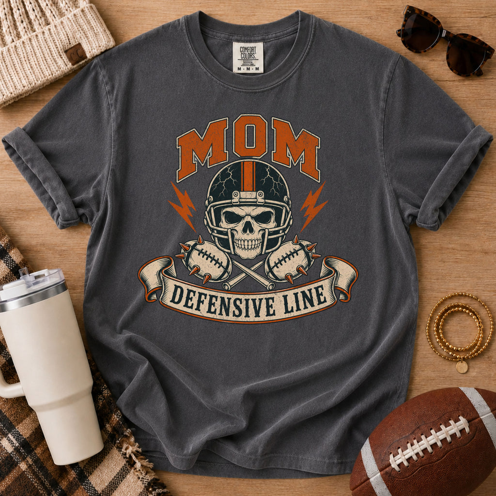 Defensive line football mom graphic T-shirt in charcoal gray, Comfort Colors Pepper, styled in a game day flat lay with a skull helmet design.