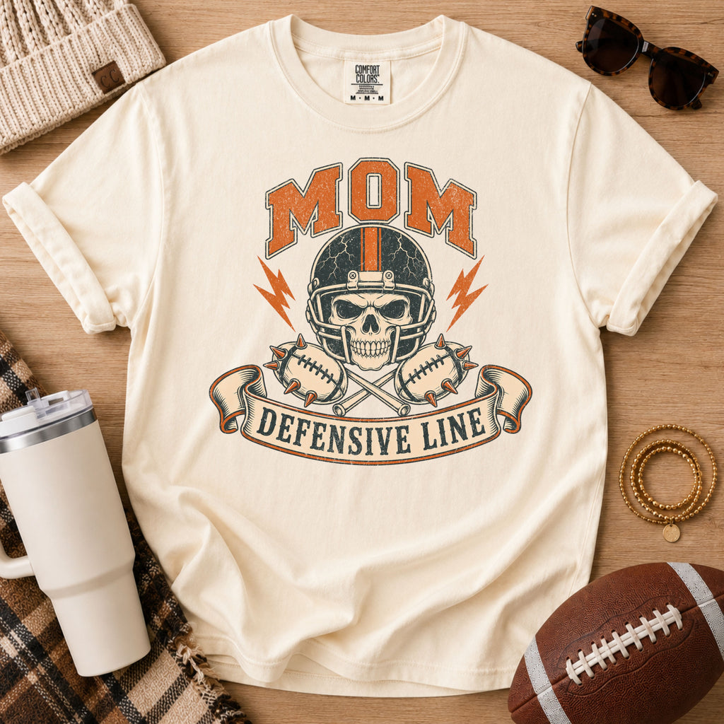 Cream football mom shirt, Comfort Colors Ivory, styled with a football and game day accessories featuring the defensive line skull graphic.