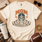 Cream football mom shirt, Comfort Colors Ivory, styled with a football and game day accessories featuring the defensive line skull graphic.