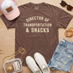 Flat lay of a distressed chocolate brown t-shirt with the slogan 'DIRECTOR OF TRANSPORTATION & SNACKS' and various casual accessories.