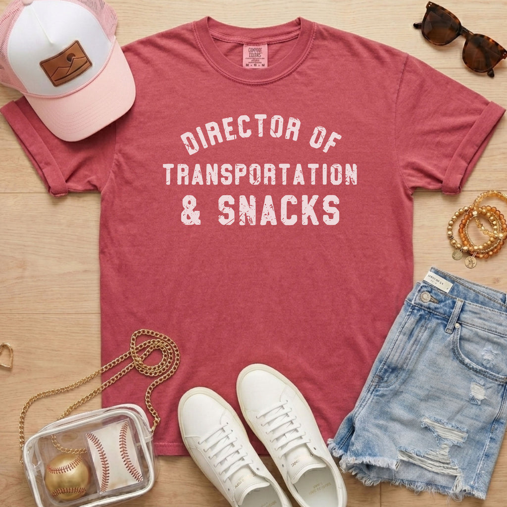 Flat lay of a distressed dusty rose t-shirt with the slogan 'DIRECTOR OF TRANSPORTATION & SNACKS' and gold accents.