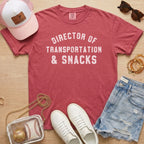 Flat lay of a distressed dusty rose t-shirt with the slogan 'DIRECTOR OF TRANSPORTATION & SNACKS' and gold accents.