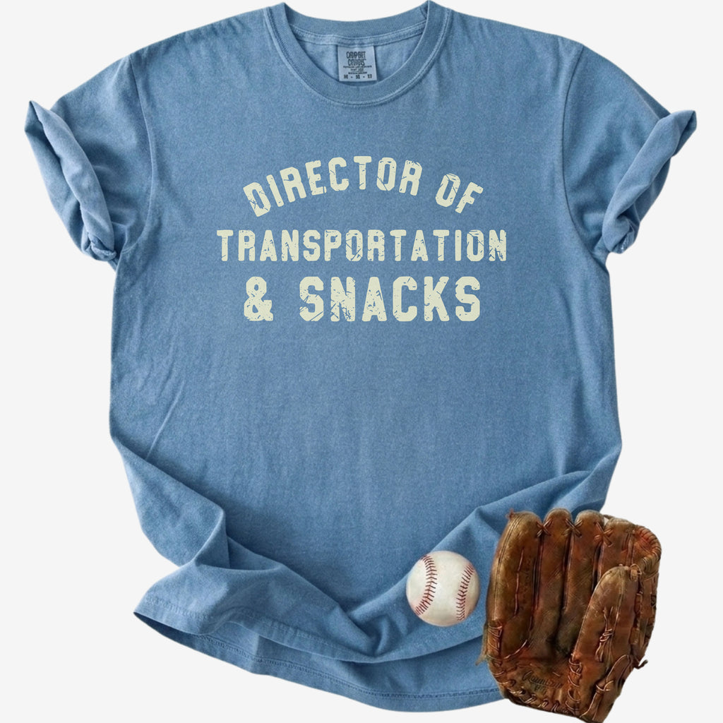 Flat lay of a distressed dusty blue t-shirt with the slogan 'DIRECTOR OF TRANSPORTATION & SNACKS' and denim shorts.