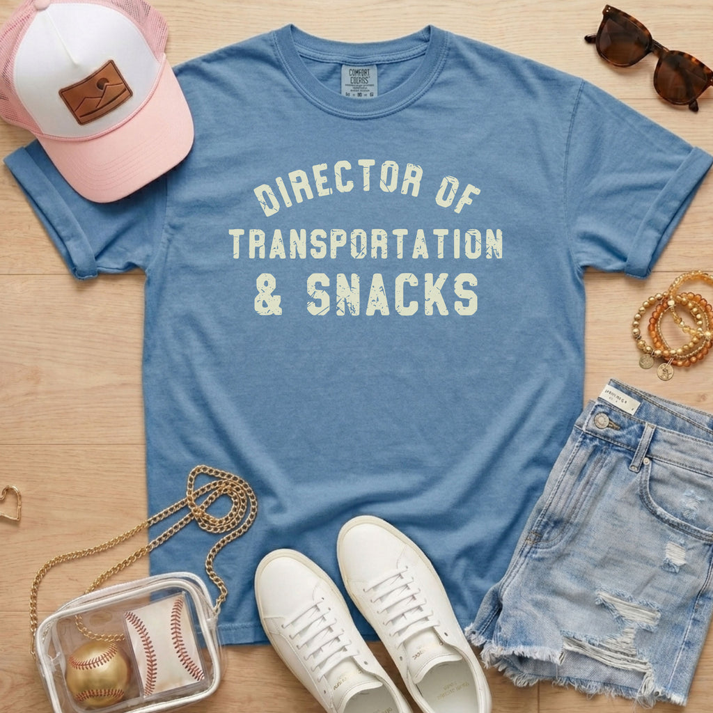 Flat lay of a distressed dusty blue t-shirt with the slogan 'DIRECTOR OF TRANSPORTATION & SNACKS' and denim shorts.