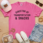 Flat lay of a distressed hot pink t-shirt with the slogan 'DIRECTOR OF TRANSPORTATION & SNACKS' and baseball accessories.