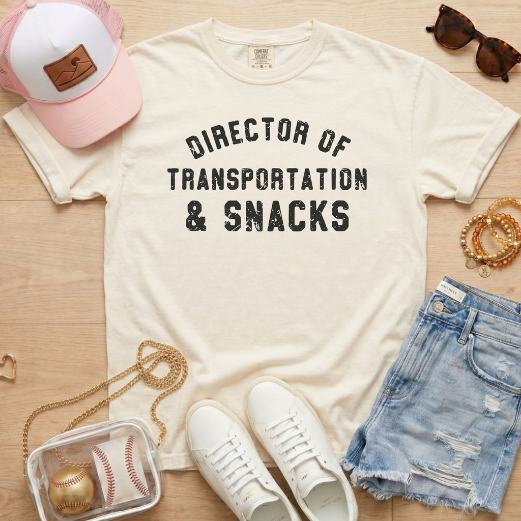 Flat lay of a distressed ivory cream t-shirt with the slogan 'DIRECTOR OF TRANSPORTATION & SNACKS' and a clear stadium bag.