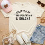 Flat lay of a distressed ivory cream t-shirt with the slogan 'DIRECTOR OF TRANSPORTATION & SNACKS' and a clear stadium bag.