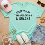 Flat lay of a distressed mint green t-shirt with the slogan 'DIRECTOR OF TRANSPORTATION & SNACKS' and tortoise sunglasses.